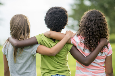 A rear view of a multi-ethnic group of elementary age girl friends are standing together with their arms around each other. 